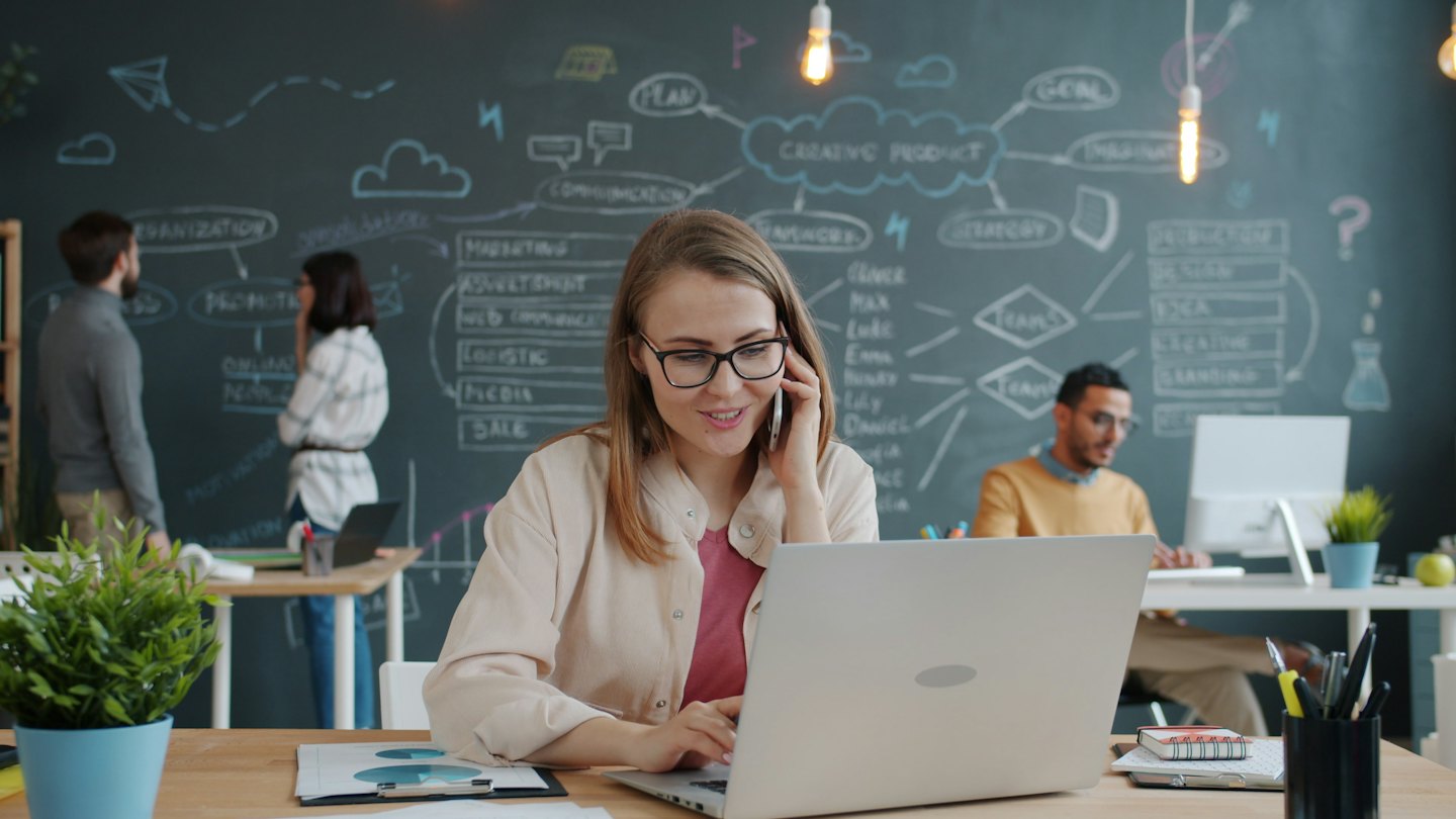 Eine Frau mit Brille lächelt, während sie im Büro an einem Laptop arbeitet und telefoniert.